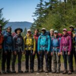 Grupa turystów na leśnej ścieżce w różnym ubraniu trekkingowym, w tle góry i niebo.