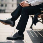 Osoba w eleganckich oksfordkach i garniturowych spodniach siedząca na ławce w miejskim otoczeniu