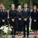 Grupa osób w czerni na cmentarzu podczas pochmurnego dnia, w tle nagrobki i białe lilie.